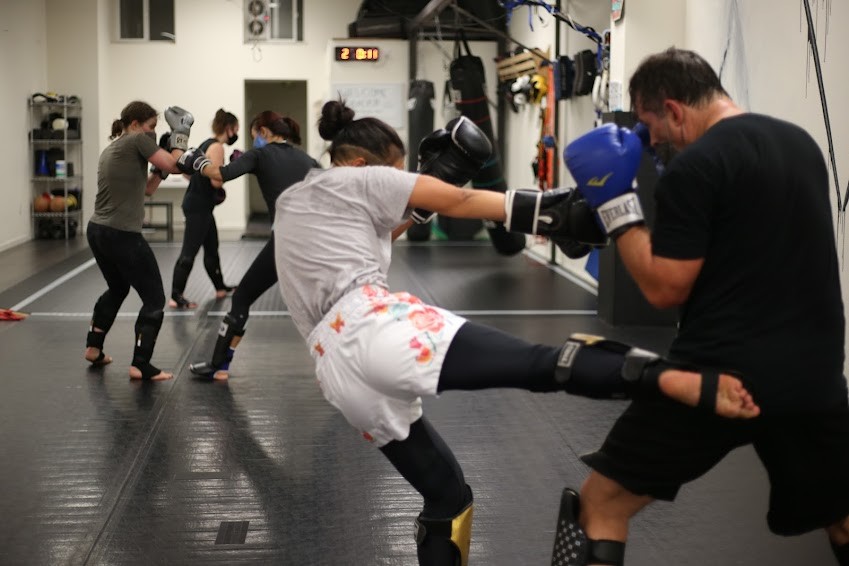 A small woman kicks the leg of a large man as other students kickbox in the background