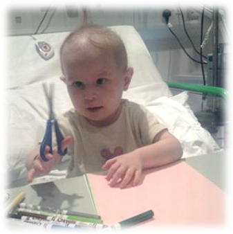 A two year old girl sits in a hospital bed with pillows and medical equipment behind her. She has little hair and her left eye is turned inward. She is holding a pair of child's scissors and there is pens and paper on the table in front of her.