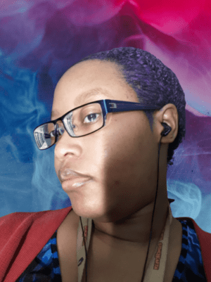 Basil Wright, a Black and Indigenous person. They have purple hair and are wearing blue glasses, a red cardigan, a blue blouse, and have black earbuds in. They stare imperiously at the camera.