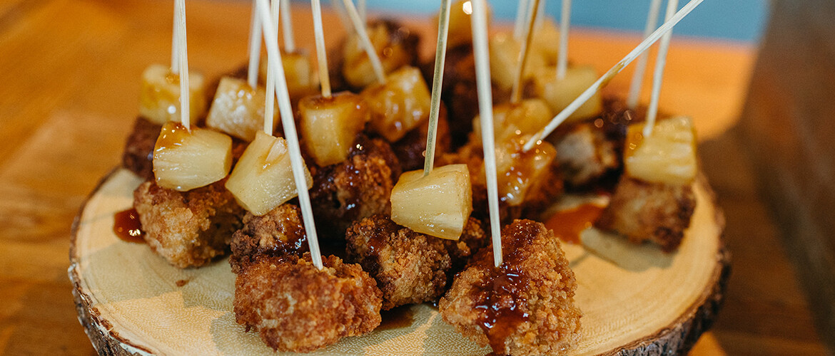 A photograph of fried chicken and pineapple skewers on a wooden tray.