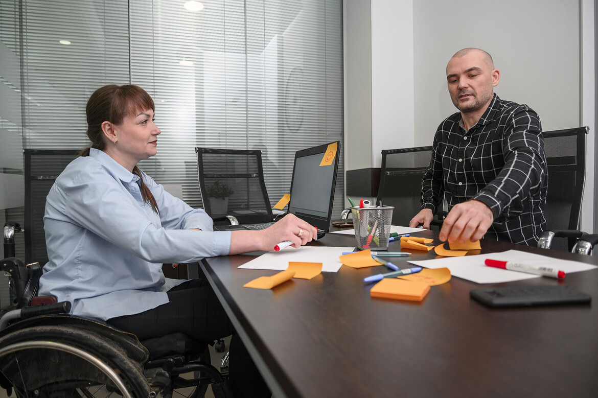 A journalist with red long red hair in a braid sits in a wheelchair on the left side of a conference table covered in sticky notes. On the right side of the table, a journalist with a shaved head navigates the table by touch.