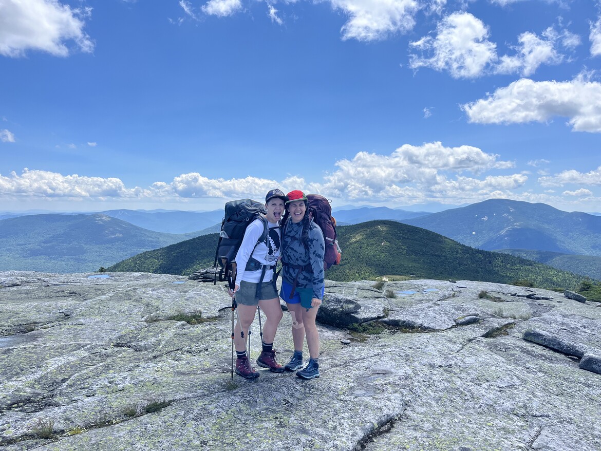 Kindling Collective co-founders on a mountain top wearing backpacking packs and holding trekking poles