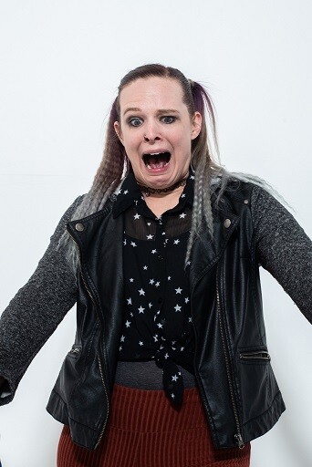 Image description: Brittany, a white person wearing a black jacket and choker necklace, makes a shocked face while looking at an invisible force beneath her.