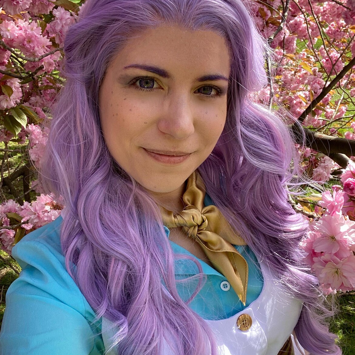 Selfie of a cosplayer dressed as Imogen Temult from Critical Role. She has a long, curly lavender wig, blue shirt, and white vest, and is surrounded by bright pink blooms in the tree behind her.