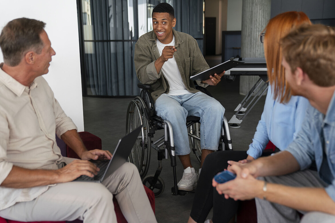 In the center of the photograph, a Black male journalist sitting in wheelchair holds a large notepad and a pen as he asks questions of two people in the right foreground of the image who face away from the camera. Another man, left, types on a laptop.