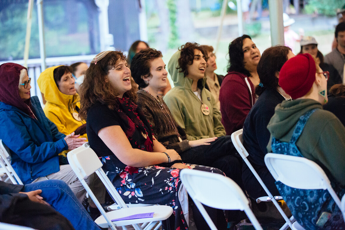 A crowd of people sitting side by side, of different ages and identities, smile while singing together.