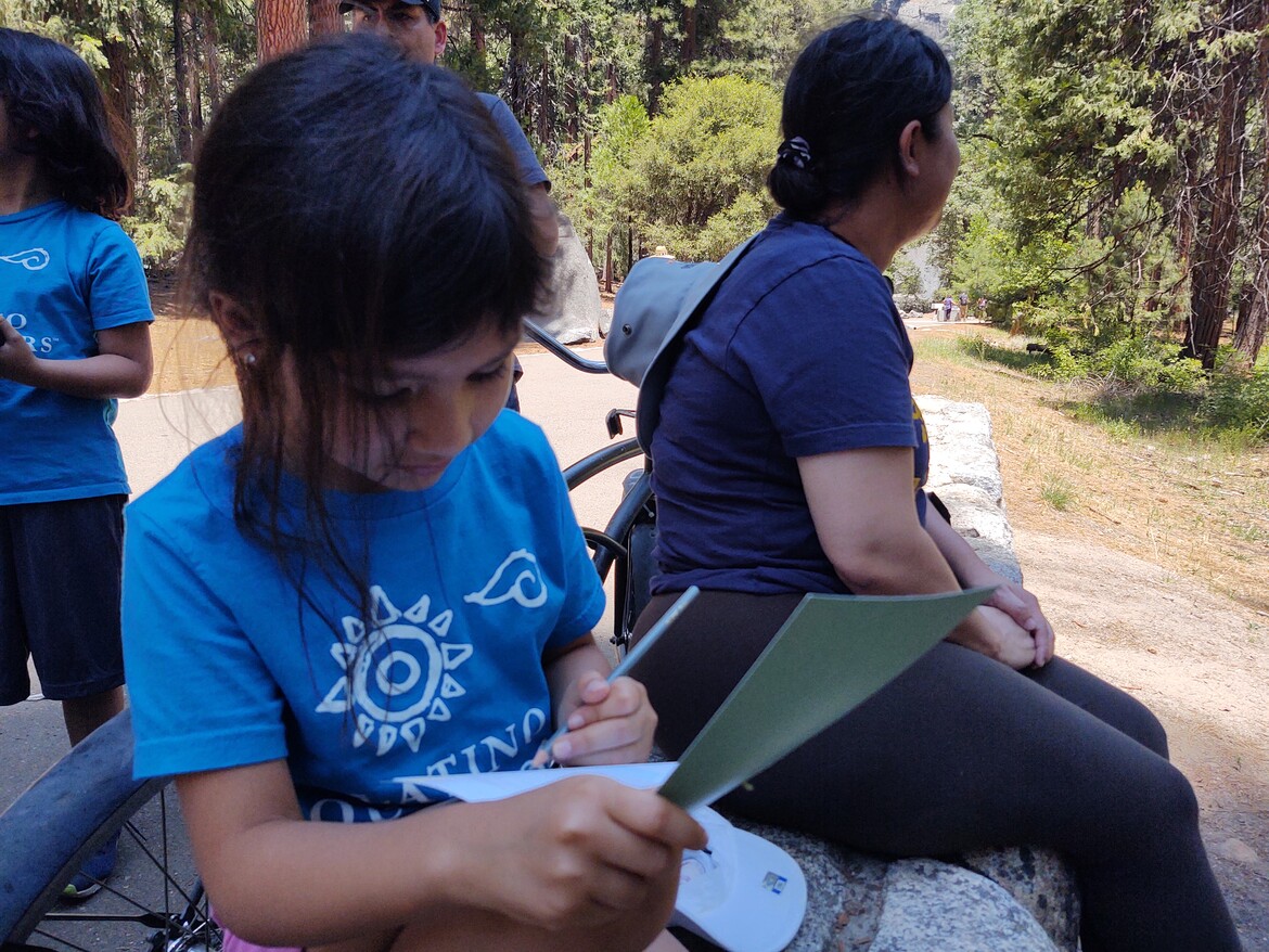 Young girl in blue Latino Outdoors T-shirt journaling while sitting on a log outdoors.